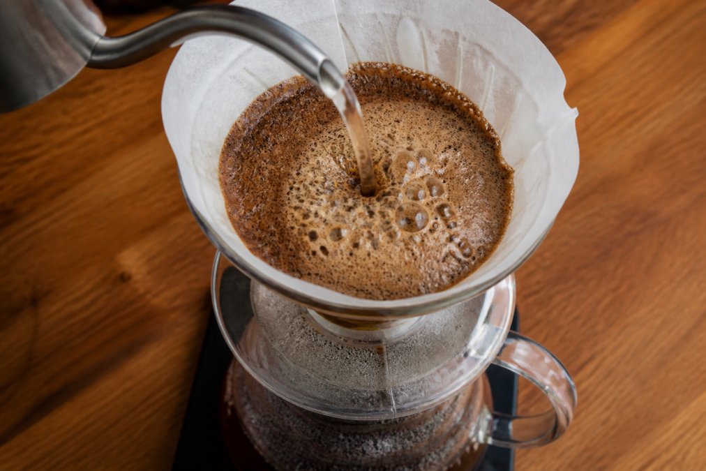 Close-up overhead shot of a pour-over coffee brewer with hot water hitting freshly ground coffee, showing thick foamy bubbles and a rising bloom in a glass carafe on a wooden kitchen counter
