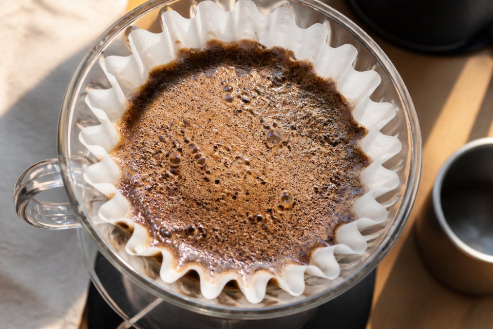 Overhead close-up of a glass pour-over brewer showing freshly ground coffee blooming with a bubbly, expanding crust after hot water was poured, warm morning light across the counter