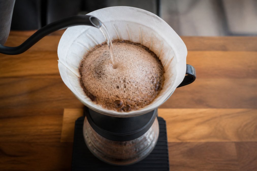 Overhead shot of hot water being poured in a slow, steady stream over dark coffee grounds in a ceramic pour-over dripper, with a large, bubbling dome of foam rising visibly from the grounds, set on a wooden counter with soft kitchen lighting