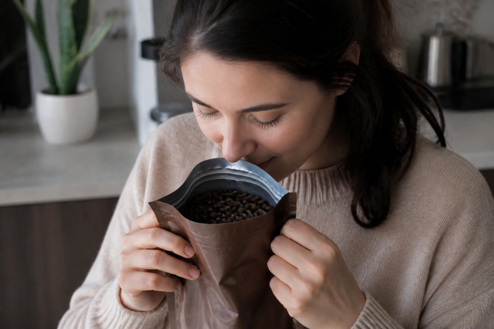 Person holding open coffee bag near their nose, inhaling deeply with eyes closed, fresh beans visibly inside the bag, natural indoor lighting