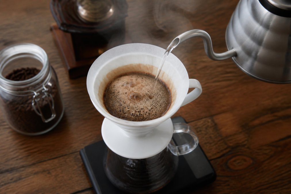 Overhead shot of a pour-over coffee dripper on a glass carafe, hot water being poured over grounds that are actively blooming and bubbling with carbon dioxide, steam rising in a warm kitchen setting