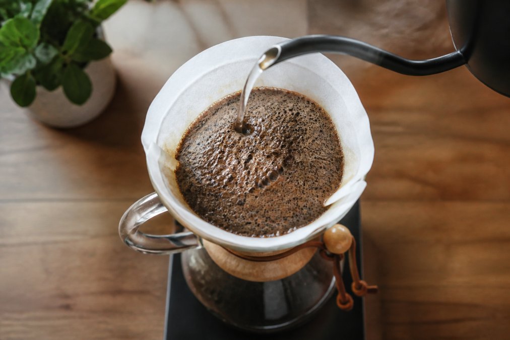 Overhead shot of a pour-over dripper with freshly ground coffee blooming, dark grounds bubbling and doming upward as hot water is poured from a gooseneck kettle, steam rising in soft morning light
