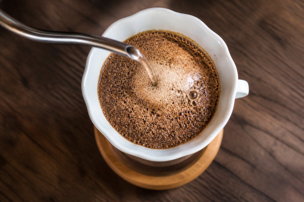Overhead shot of a slow, steady pour of hot water from a gooseneck kettle over coffee grounds in a ceramic pour-over dripper, with a visible dome of foam and CO2 bubbles rising from the dark coffee bed on a wooden surface