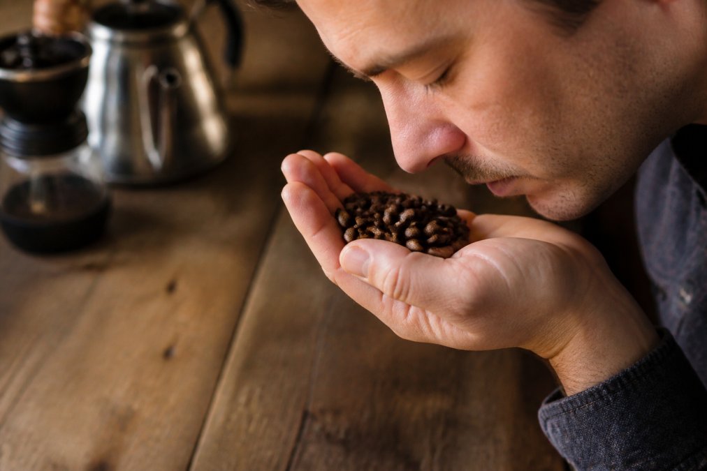Close-up of a person's hand holding a small handful of dark roasted coffee beans near their nose, eyes closed, performing a smell test over a rustic wooden table in warm kitchen lighting
