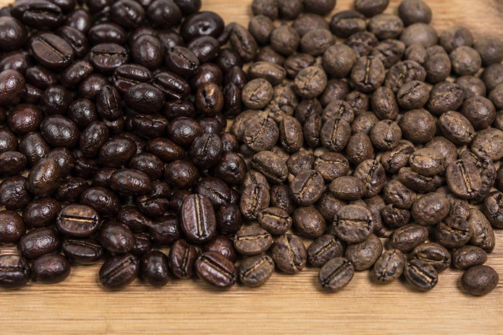 Close-up of fresh dark roast coffee beans with glossy oil sheen next to older beans with dull, chalky surface showing visible color fading and uneven texture