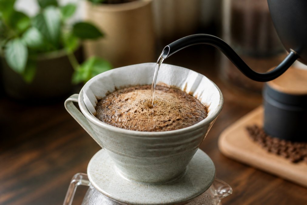Close-up of hot water being poured from a gooseneck kettle over dark coffee grounds in a ceramic pour-over dripper, with a visible dome of foam and bubbles rising from the grounds in warm kitchen lighting