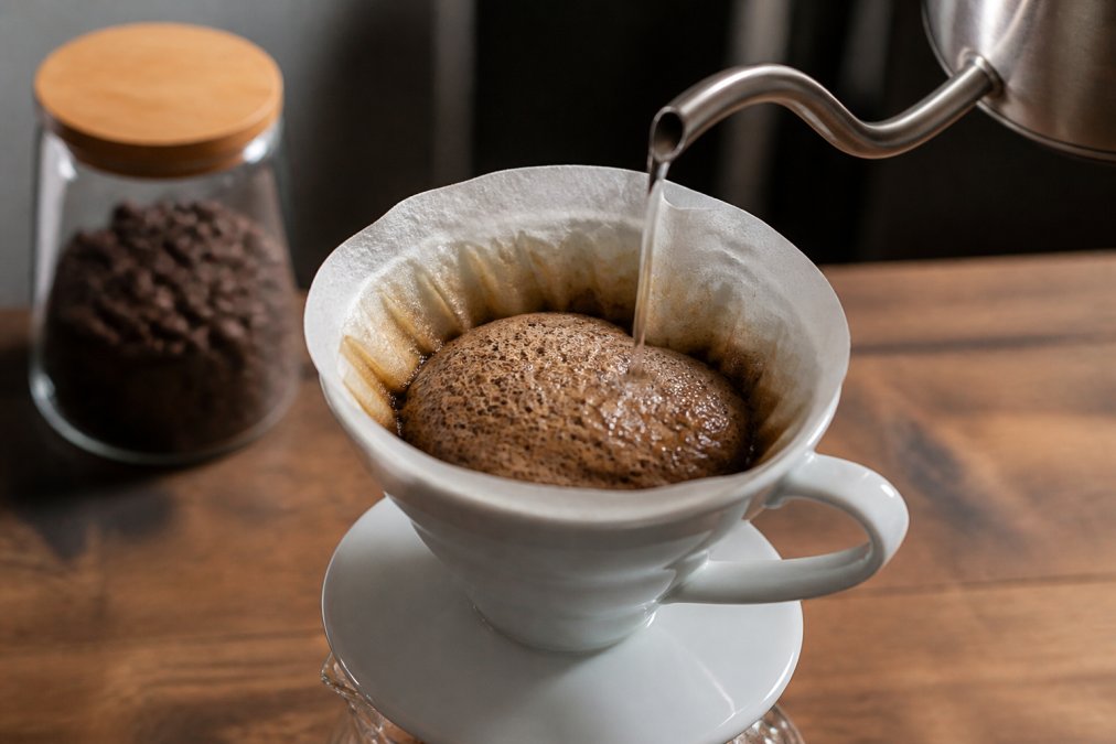 Close-up of hot water being poured from a gooseneck kettle over coffee grounds in a ceramic pour-over dripper, with a visible dome of CO2 bubbles blooming upward from the dark, saturated grounds on a wooden kitchen counter