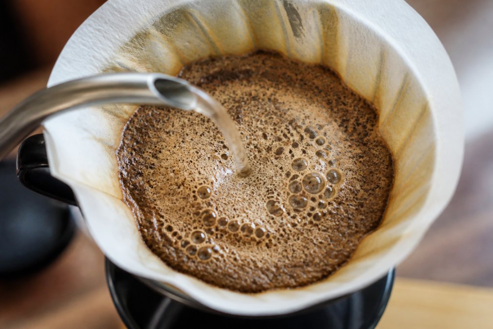Close-up overhead shot of hot water being poured over ground coffee in a pour-over dripper, with foamy bubbles and blooming CO2 visible on the surface