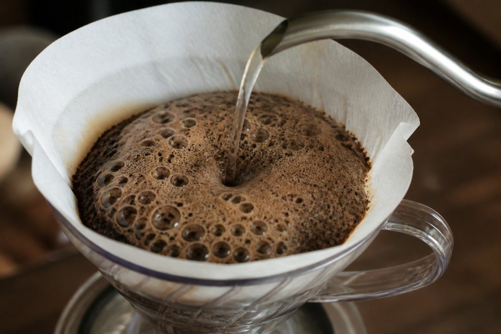 Close-up of a pour-over coffee brewer with hot water hitting coffee grounds, showing a thick bubbling bloom of foam rising in the cone