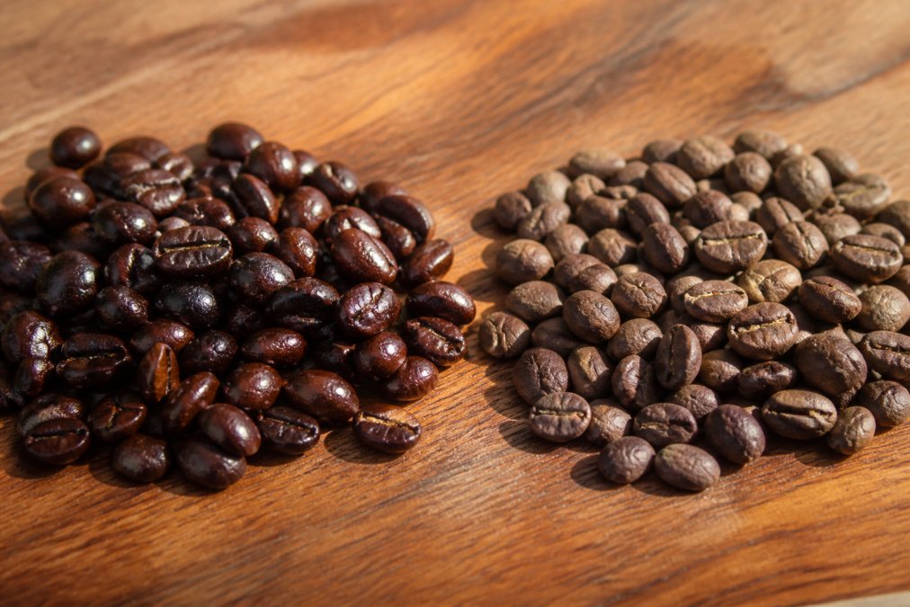 Close-up of two piles of whole coffee beans side by side in natural daylight, glossy, richly-colored fresh beans with visible shine next to dull, flat-looking stale beans with no luster