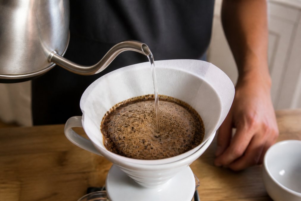 Hands pouring hot water from a kettle over finely ground coffee in a white filter cone, with visible CO2 bubbles and foam rising from the coffee grounds in a bright kitchen setting