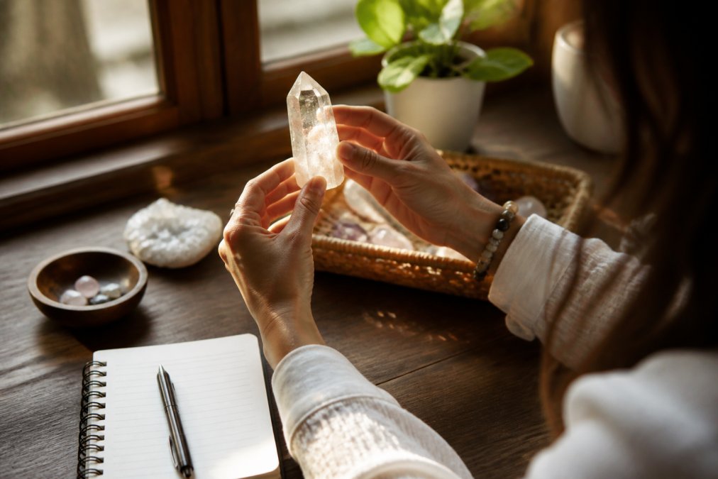 A woman's hands holding a clear quartz crystal up to warm window light, examining it closely with a small notebook and pen resting on a wooden table nearby