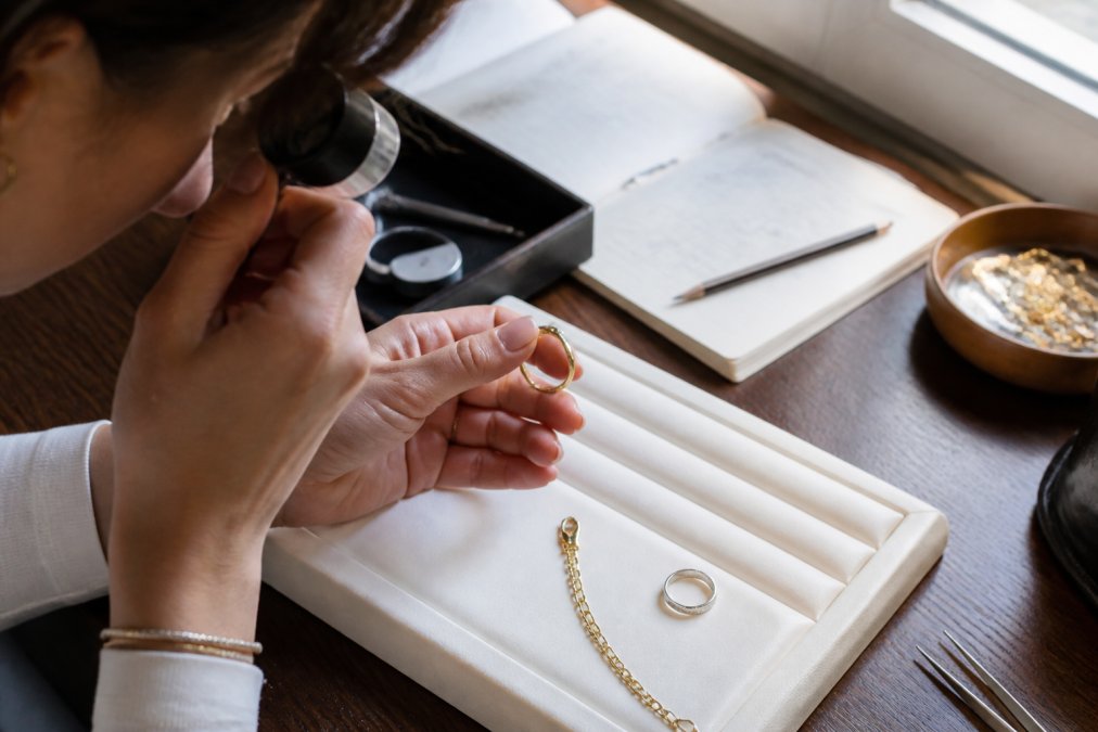 Close-up of a person using a magnifying loupe to examine small hallmark stamps on the inside of a gold ring, the ring resting on a white velvet surface under soft natural window light