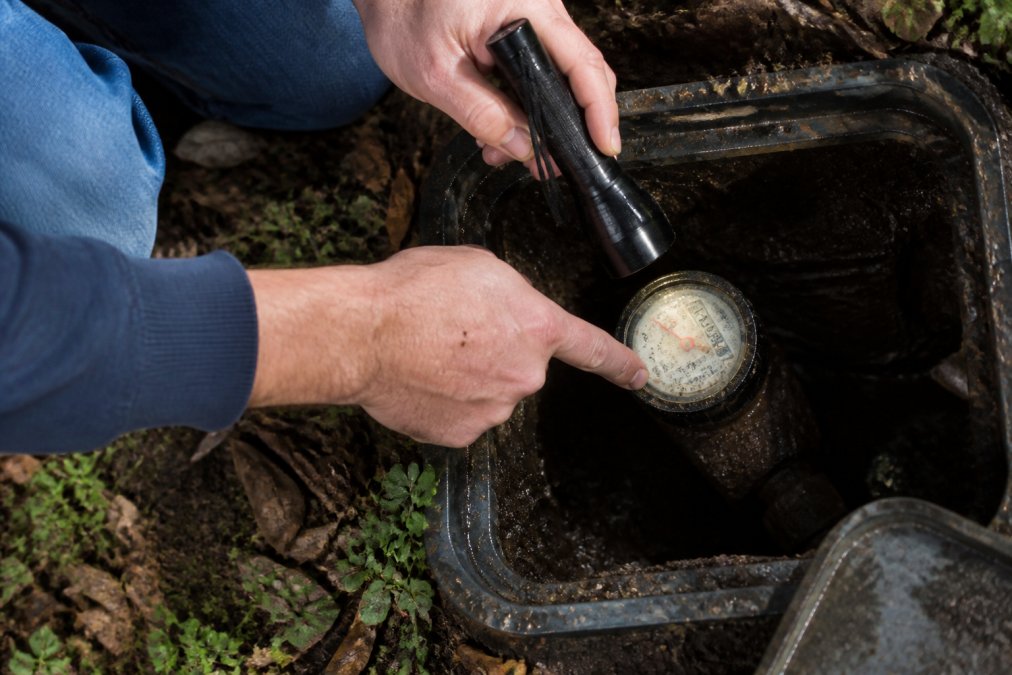 Homeowner's hands checking a water meter dial in the yard, pointing to the meter reading with a flashlight to identify any movement