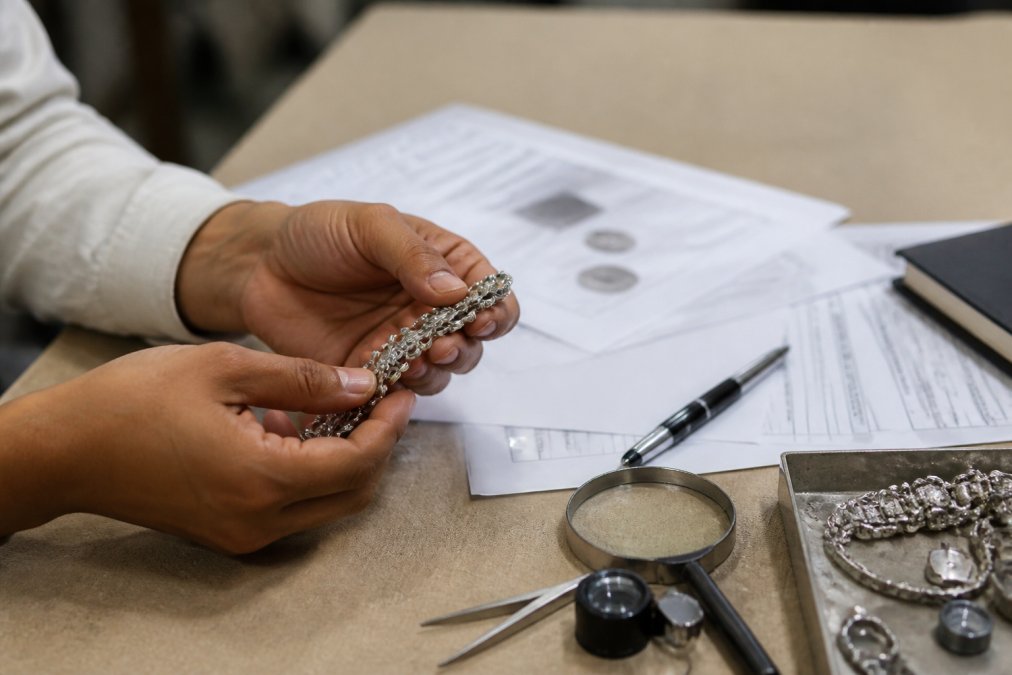 Close-up of hands holding silver jewellery piece with certification documents and supply chain traceability papers spread on a neutral surface, warm office lighting