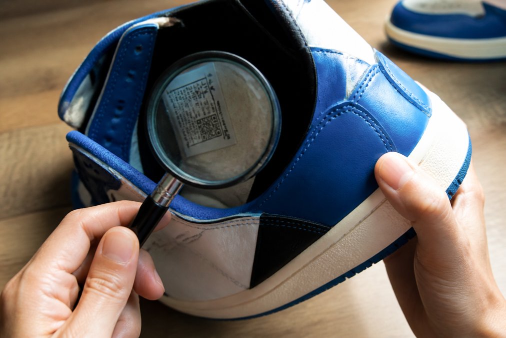 Close-up of hands holding a sneaker under bright light, examining the stitching and size tag with a magnifying glass on a clean table