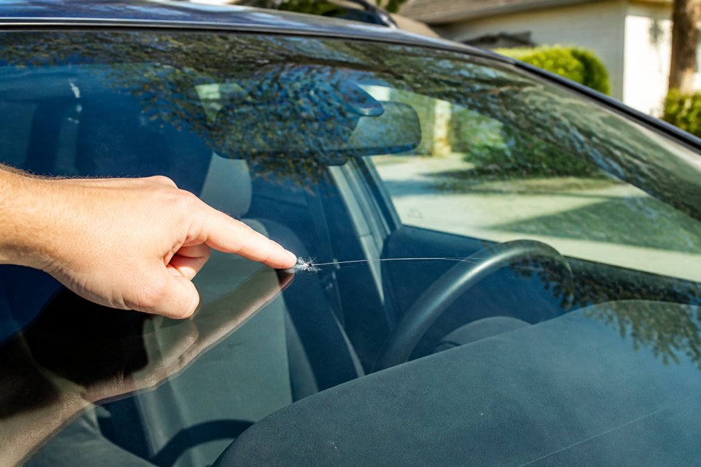 Close-up of a car windshield with a visible chip and crack, a person's hand pointing at the damage to show size and location, parked in a driveway with daylight reflecting off the glass