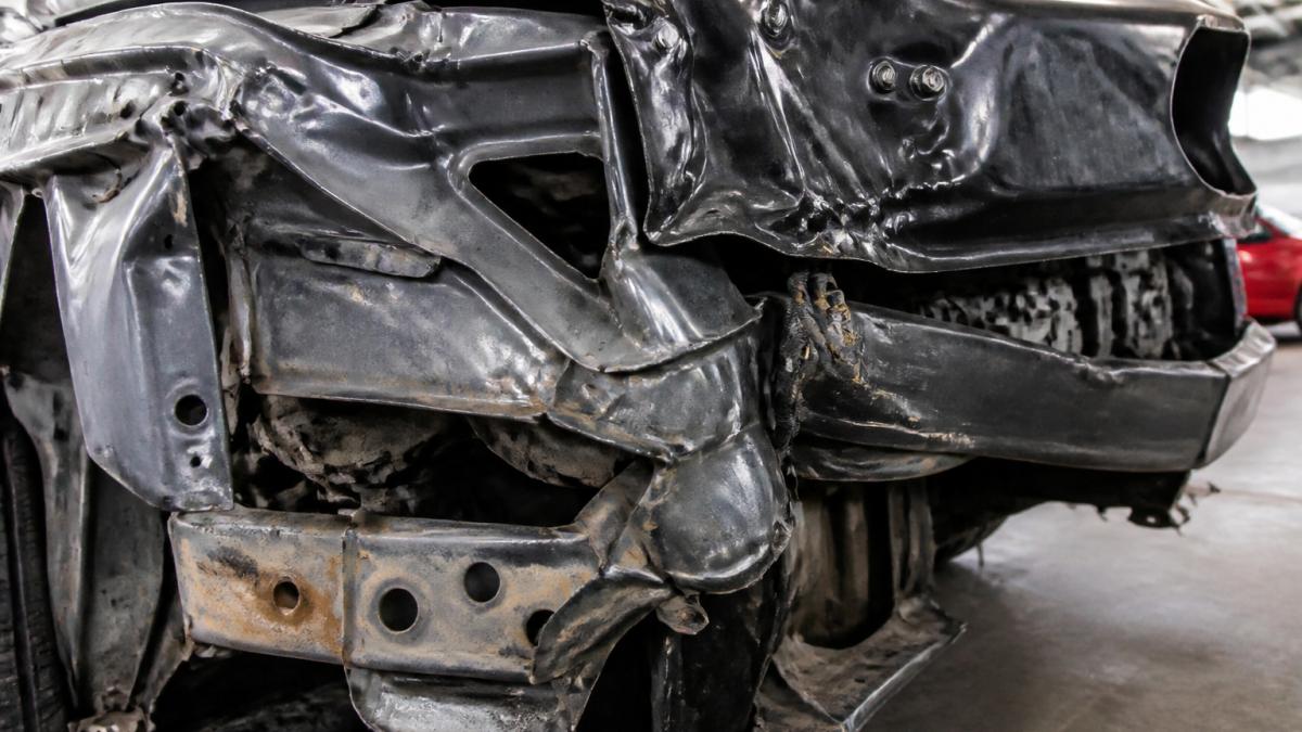 Close-up of severely crumpled vehicle frame showing bent metal, twisted structural components, and deformation from collision impact in repair shop lighting