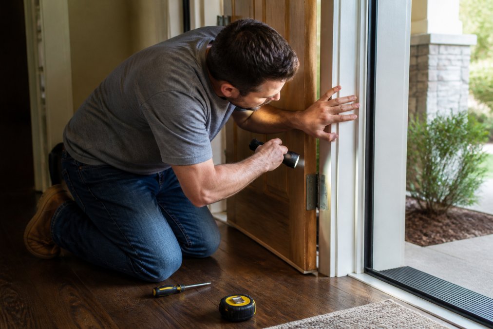 A homeowner kneeling at an open front door, running a hand along the door jamb and hinge side while holding a flashlight, with a screwdriver and tape measure resting on the floor nearby