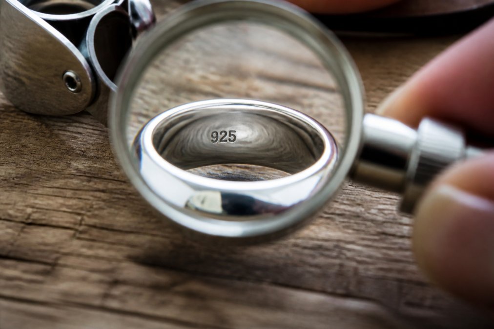 Close-up of a magnifying glass examining hallmark stamps on the back of a silver ring, showing clear 925 marking on polished metal surface with natural lighting