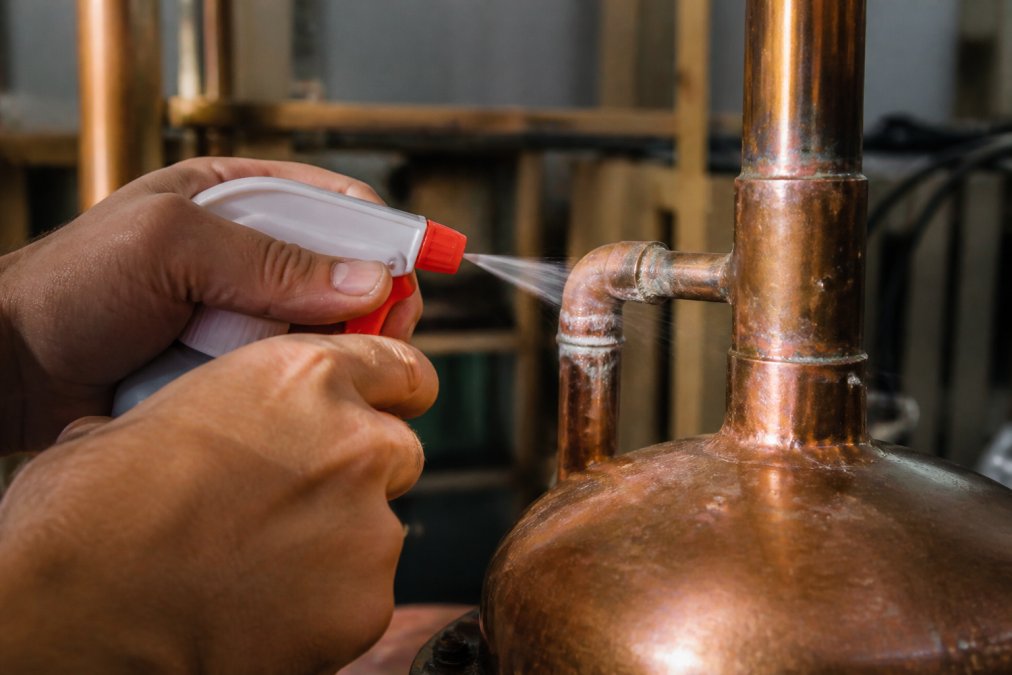Hands spraying soapy water on copper joints of a still for leaks as bubbles form at the connection point