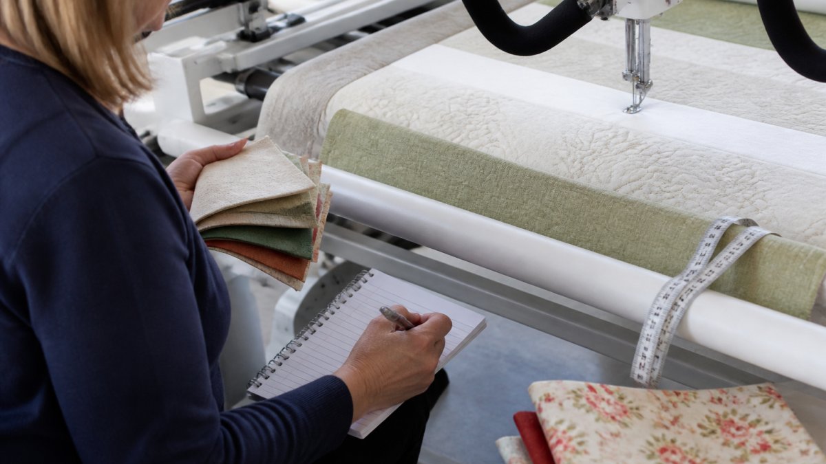 Quilter's hands holding fabric samples and taking notes while examining longarm quilting machine stitching detail with measuring tape visible on quilting frame
