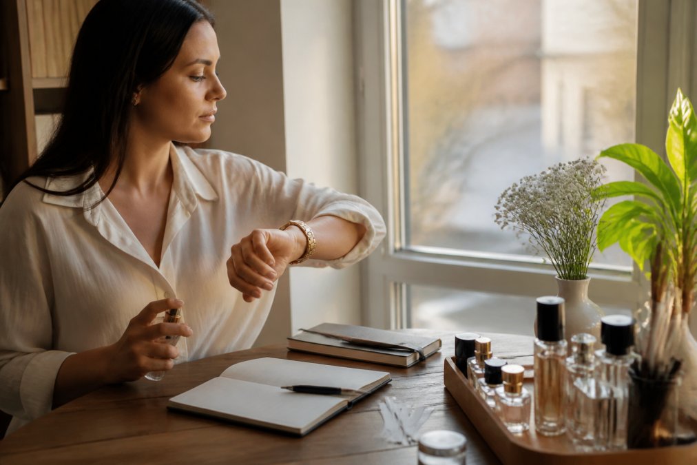 A person checking their watch while seated by a window with natural afternoon light, wearing a fragrance on their wrist, with a notebook nearby for tracking scent evolution throughout the day