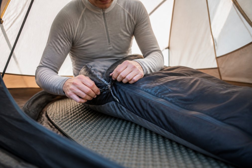 Close-up of a person in a grey thermal base layer top zipping up a dark sleeping bag inside a tent, with a foam sleeping pad visible underneath and soft morning light filtering through the tent fabric