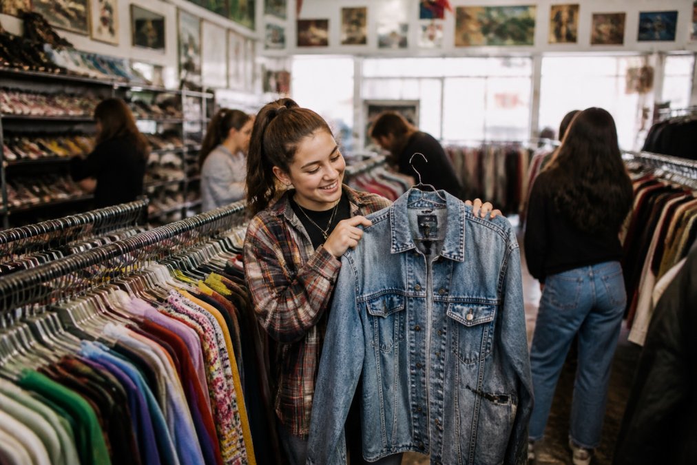 A teenager browsing through racks of colorful secondhand clothing in a bright thrift store, holding up a vintage denim jacket with a smile, other shoppers in the background