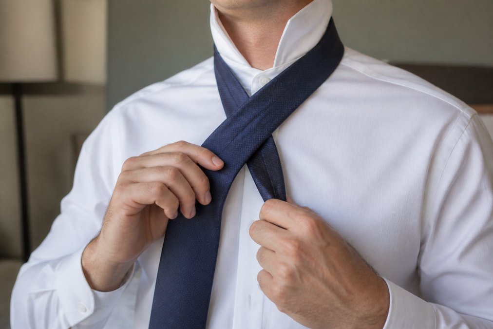 Close-up of a man's hands in the middle of tying a silk necktie into a four-in-hand knot, with the wide end crossing over the narrow end, crisp white shirt collar visible, soft natural light