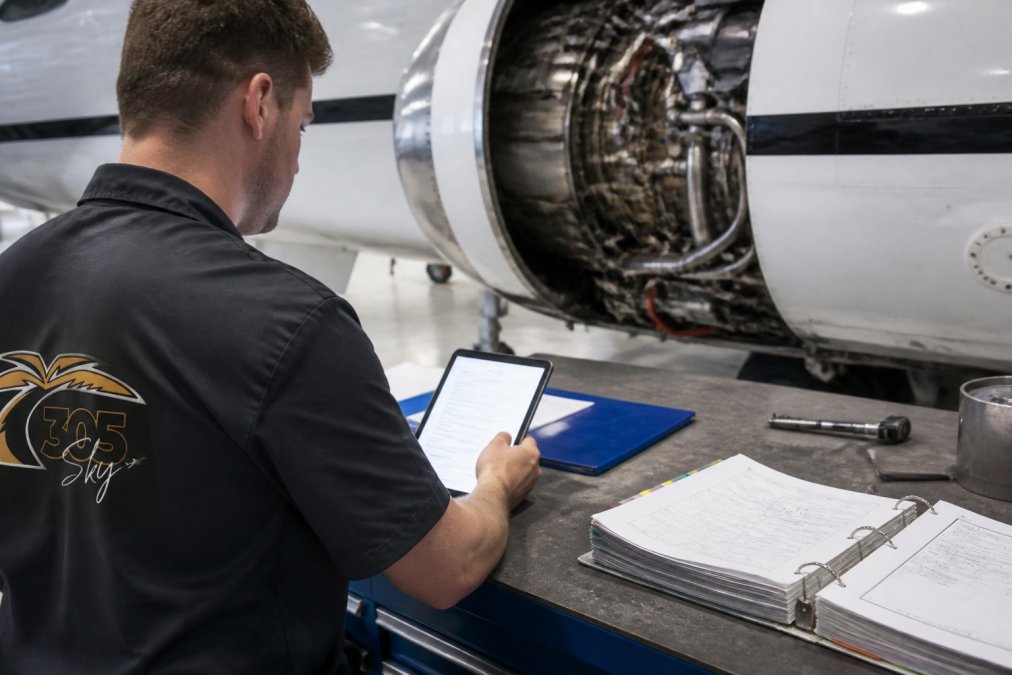 Aircraft maintenance technician reviewing engine time data on a tablet or laptop screen in a maintenance hangar, with maintenance records and technical documentation visible on the workbench