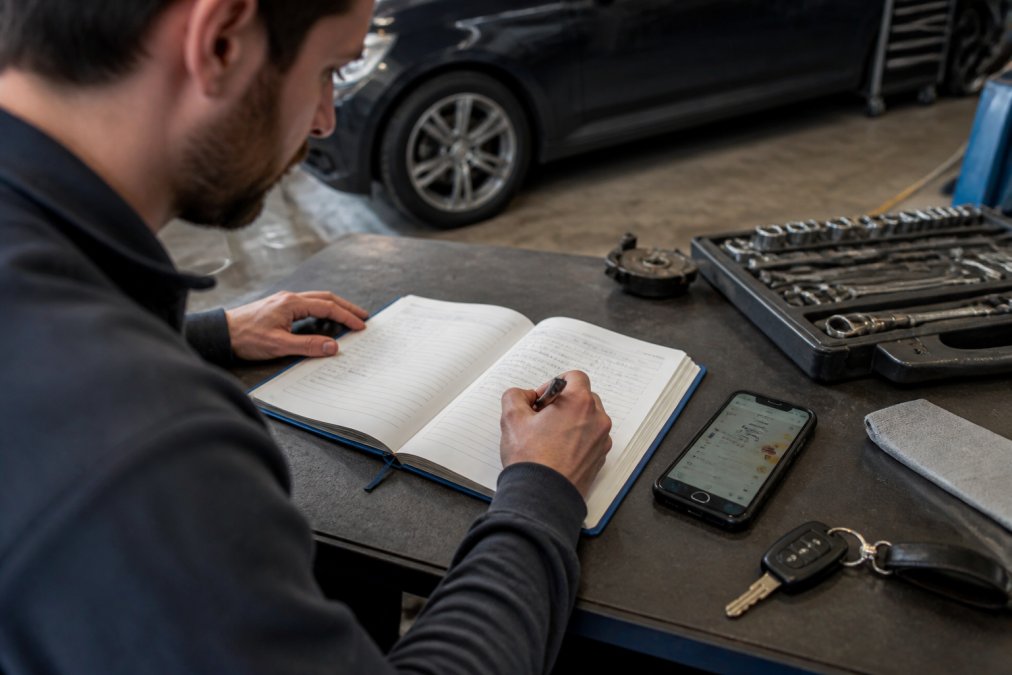 Person recording service dates and mileage in a logbook to track car maintenance alongside a digital app.
