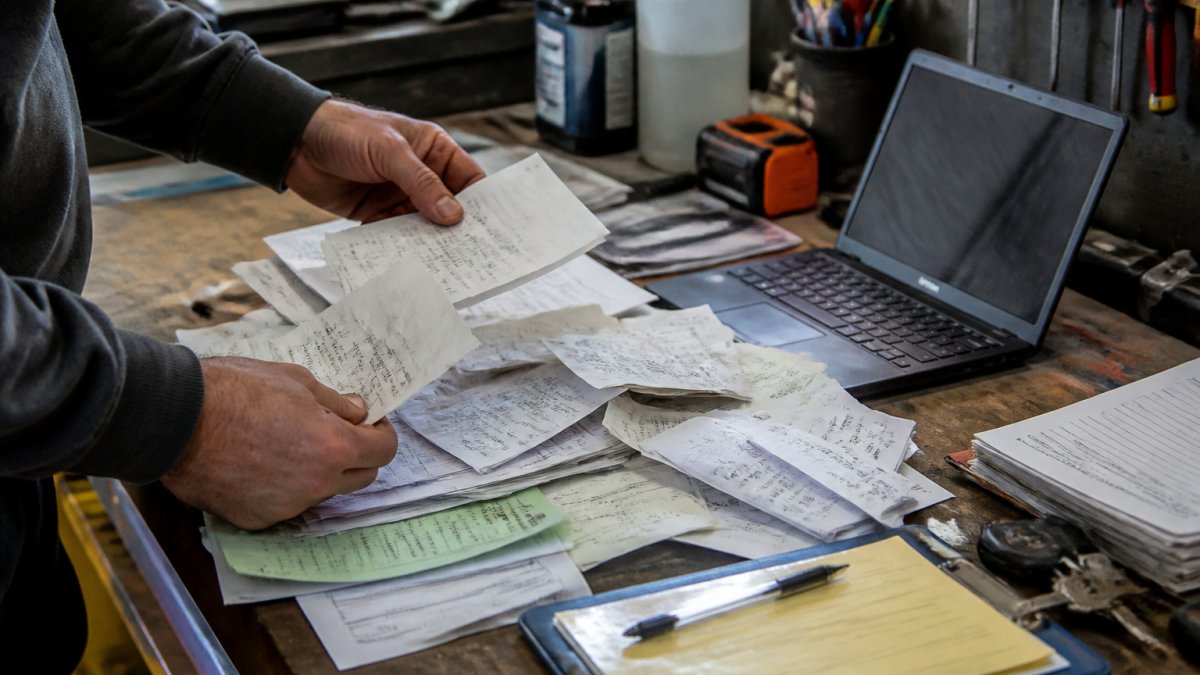 A car owner's hands sorting through a messy pile of old receipts, bank statements, and service invoices on a garage workbench, with a laptop open to a digital logbook in the background