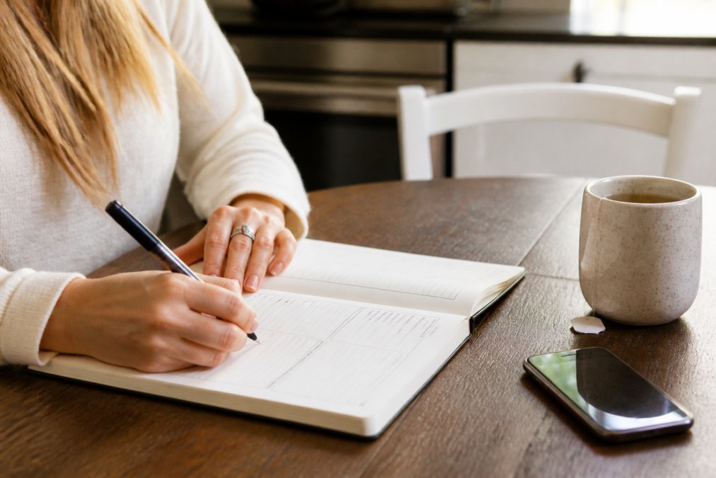 Close-up of a woman's hands writing in a paper health journal at a kitchen table, with a mug of tea and a smartphone nearby in soft natural light