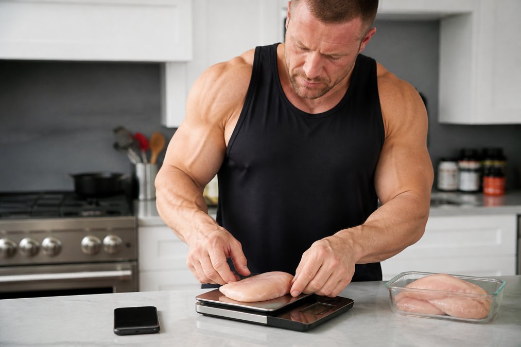 A close-up of a person's hands placing a chicken breast on a digital food scale in a bright, clean kitchen. A smartphone with a tracking app is visible nearby.