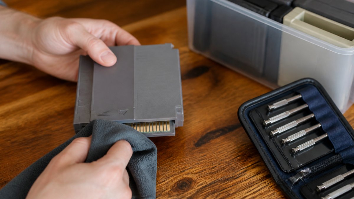 Close-up of hands using a microfiber cloth to gently clean the gold contacts of a retro game cartridge on a wooden table, with a small screwdriver kit and a labeled storage bin nearby