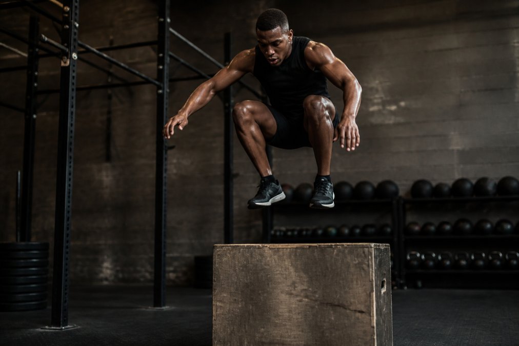 A determined athlete performing explosive box jumps in a gritty, dimly lit gym with sweat visible, wearing dark athletic apparel, focused expression