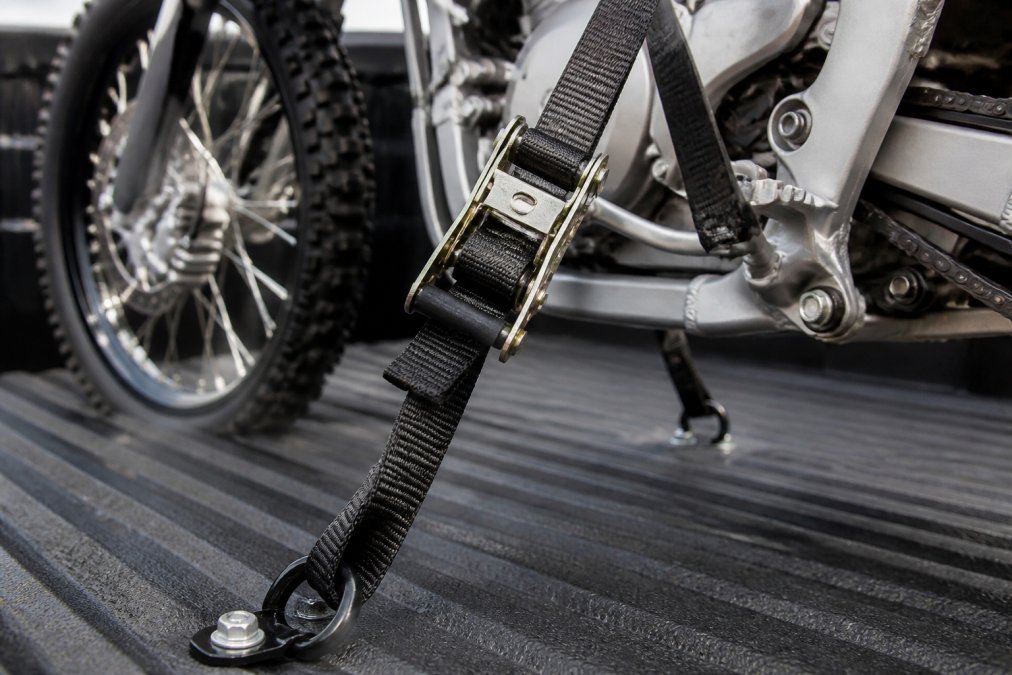 Close-up of heavy-duty ratchet straps tightly securing a dirt bike's frame to a truck bed, showing the strap routed through the frame and tightened with visible tension, metal attachment points visible