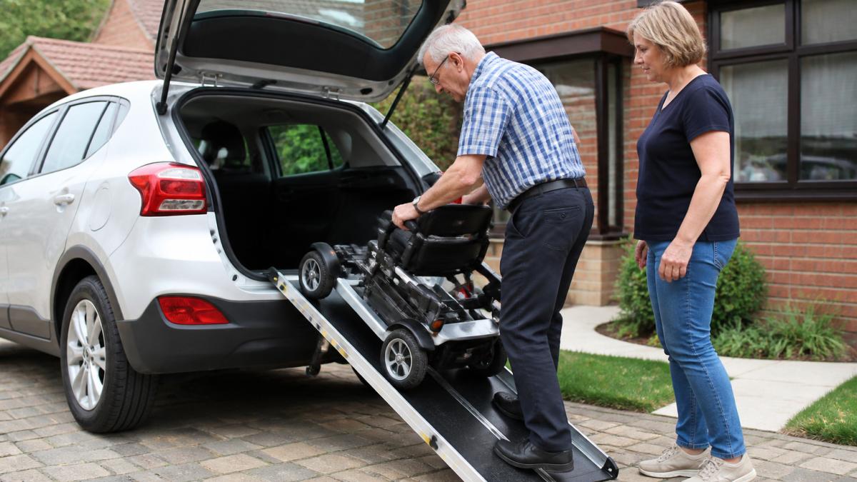 Caregiver assisting someone as they roll a folded mobility scooter up a ramp into a car boot