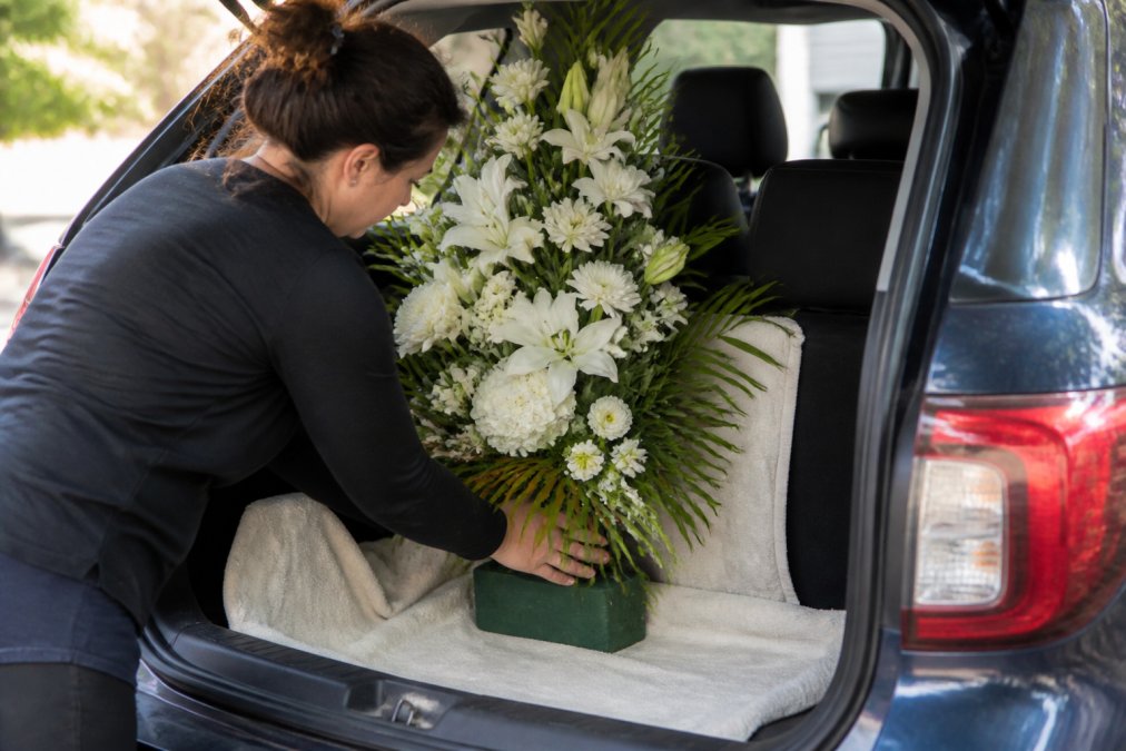 A person carefully placing a large standing spray into the back of an SUV, using towels to cushion the base and secure it against the seat. The arrangement is upright and stable, with floral foam visible at the base. Soft daylight, respectful and calm mood.