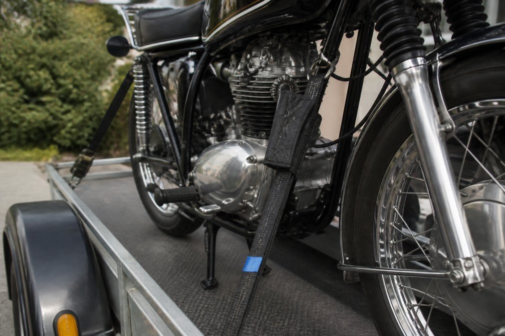 Close-up of soft padded tie-down straps properly secured to a vintage motorcycle frame on a transport trailer, showing correct anchor point placement at the main frame tubes with the strap running parallel to the bike's centerline