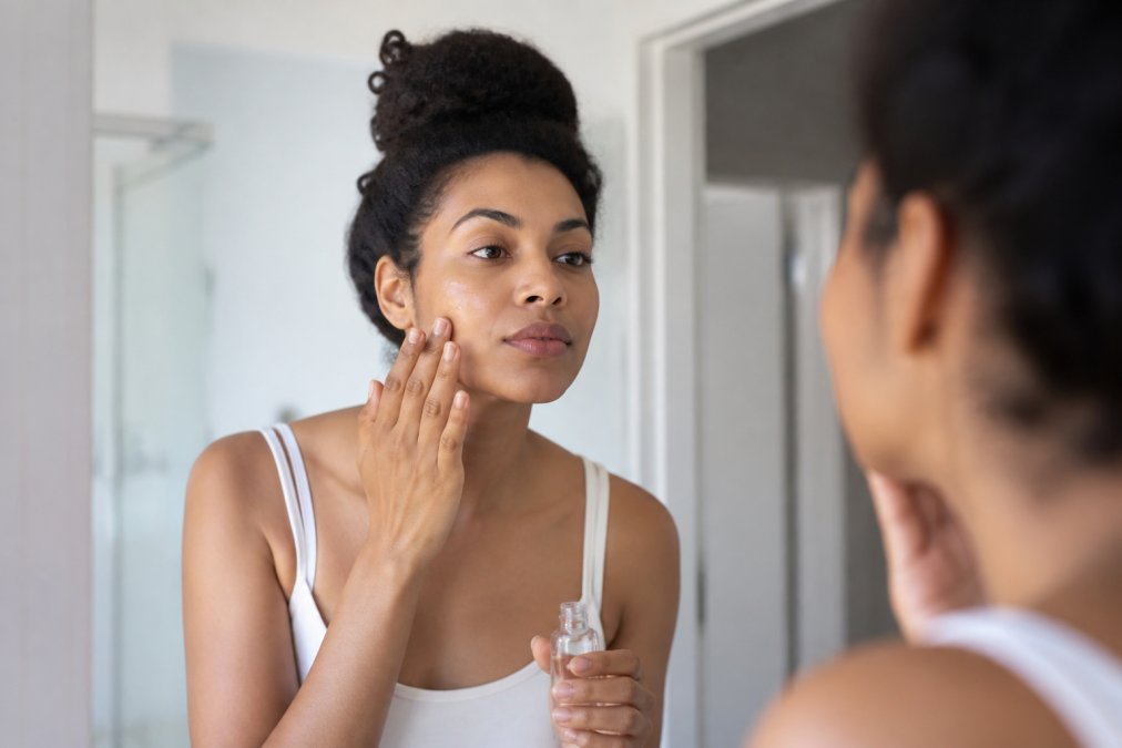 Close-up of a woman with medium-brown skin applying a clear serum to her cheek with her fingertips in a bright, clean bathroom, looking at her reflection in the mirror with a calm expression