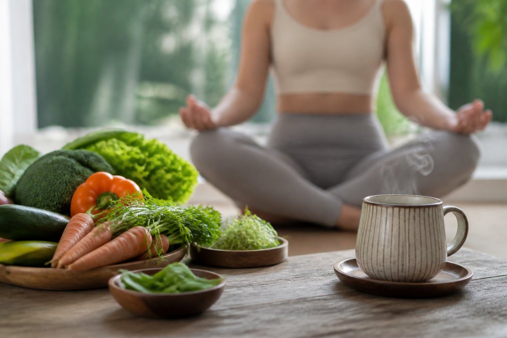 Wellness-focused scene showing fresh organic vegetables, herbal tea steaming in a ceramic cup, and person in peaceful meditation posture in bright, naturally-lit calm environment with soft natural window lighting