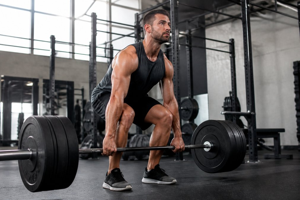 A fit man performing a barbell deadlift in a modern gym setting with proper form, showing muscular engagement through back, legs, and core, with focused expression and controlled movement