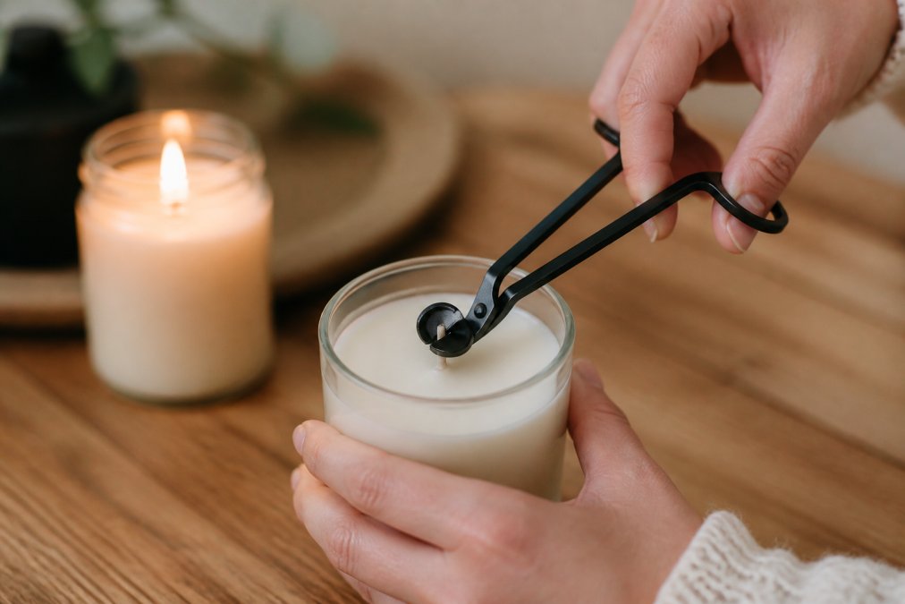 Close-up of hands using a candle wick trimmer to cut a wick to 1/4 inch length above the wax pool, with a lit candle glowing softly in the background on a wooden table