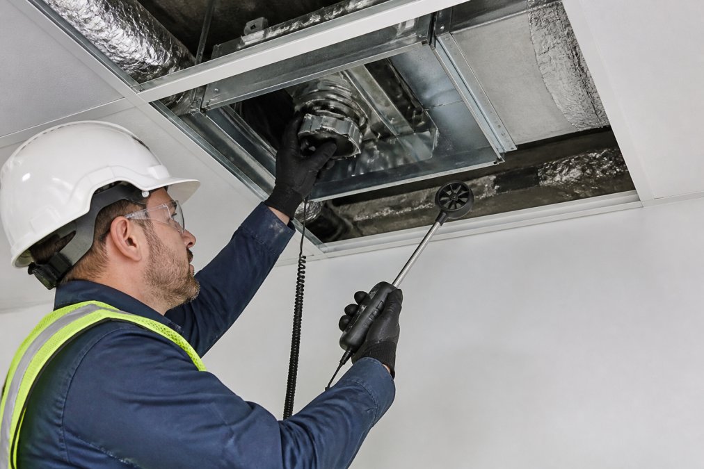 Facilities engineer in safety gear adjusting VAV box controls and checking airflow velocity with handheld instrument at a cleanroom ceiling plenum access point, with visible ductwork and damper mechanisms