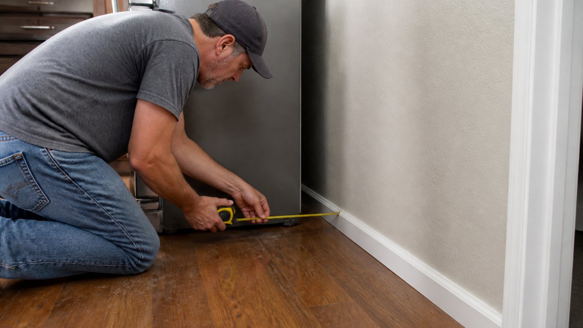 A homeowner pulling a refrigerator away from the kitchen wall to reveal the space behind it, with a tape measure in hand checking the gap near the baseboard