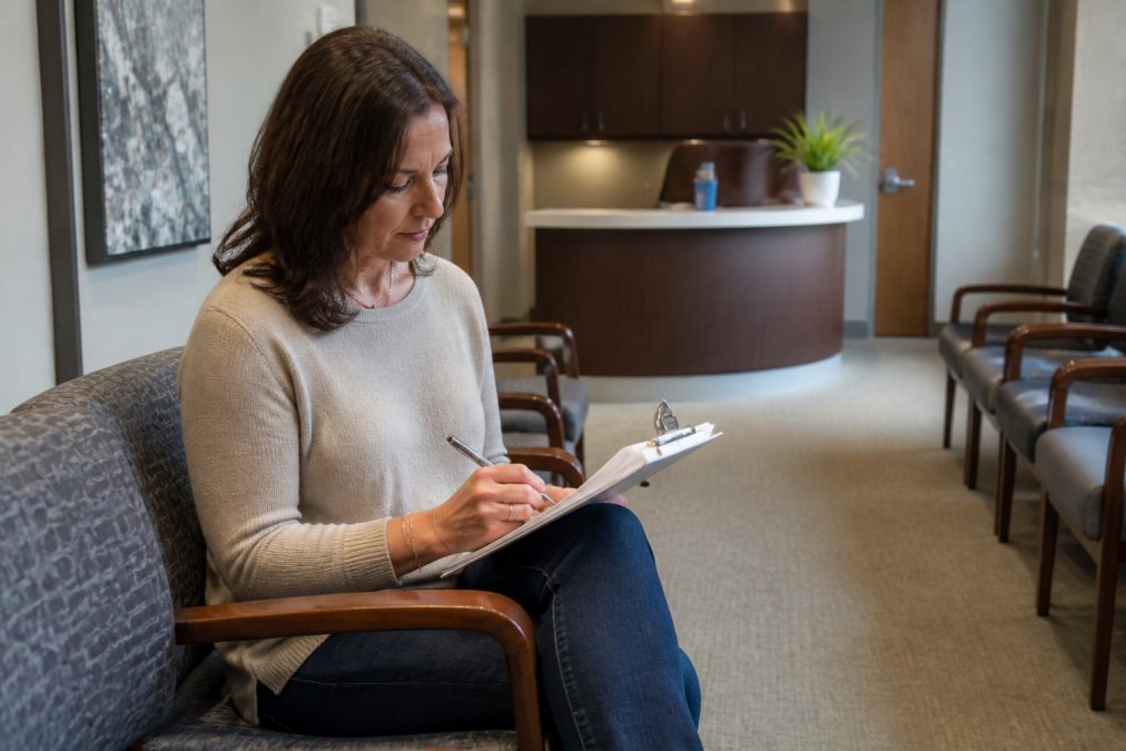 Woman in a medical office waiting room reviewing notes on a clipboard, looking prepared and engaged