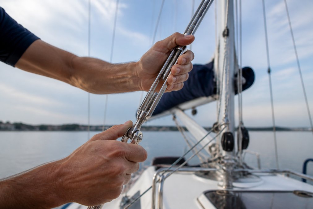 Professional sailor's hands adjusting a chrome turnbuckle on standing rigging with the mast and shrouds visible in background during calm conditions
