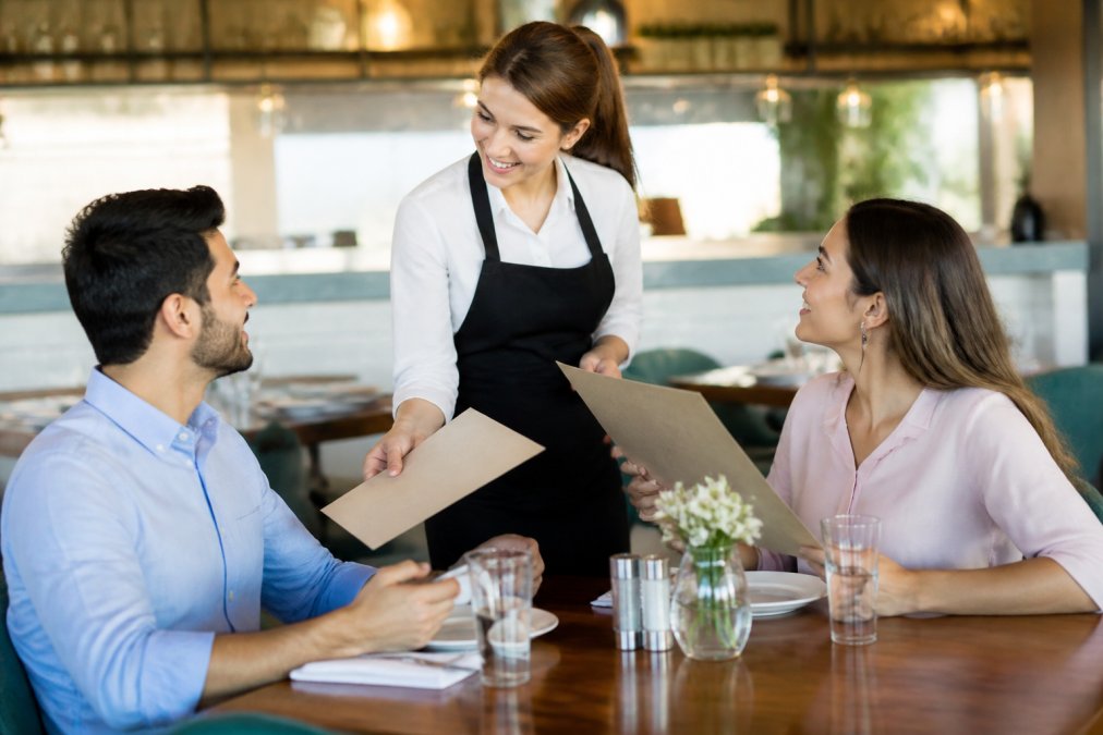 A friendly server warmly greeting a couple seated at a restaurant table, handing them menus with a genuine smile in a cozy, well-lit dining room